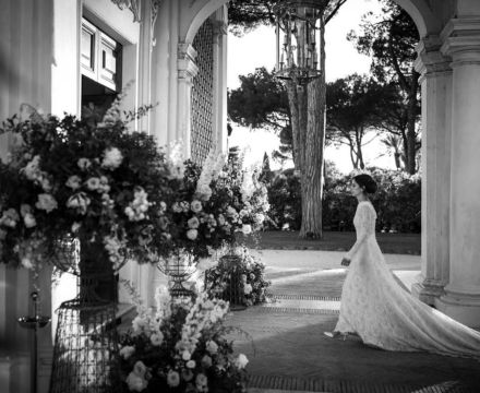 Sposa Il Giardino Fiorito delle Spose in abito in pizzo con coda lunga mentre attraversa il portico di una villa durante il matrimonio